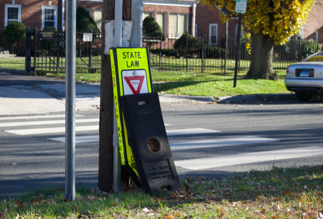 A broken yield-to-pedestrian sign on side of road by a high school. It was reported broken several months before picture taken.