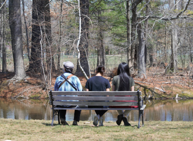Three people sitting on a bench in the park by a pond