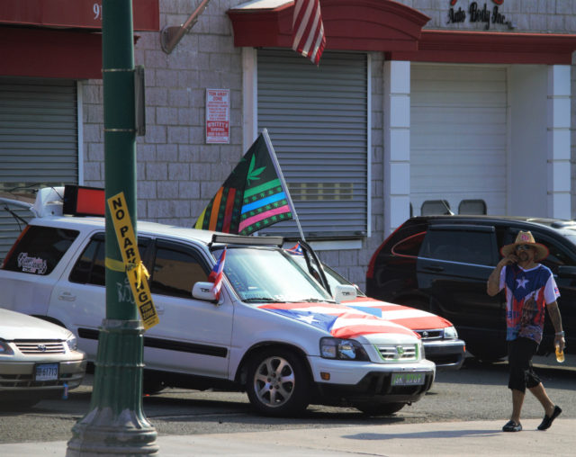 Person wearing Puerto Rican flag shirt and straw hat walking by a car that is covered in a Puerto Rican flag and flying a rainbow flag with marijuana leaf