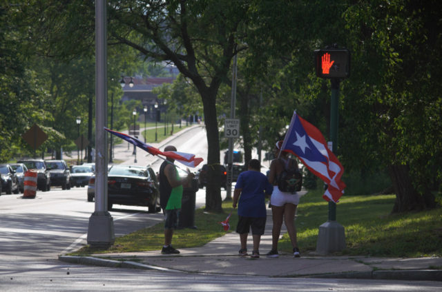 People standing on sidewalk near Pope Park, holding large Puerto Rican flags 
